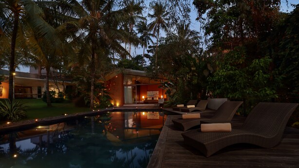 A pool area with lounge chairs and a wooden deck, surrounded by palm trees and a building with warm lighting. - Umah Tampih Villa (Ubud)