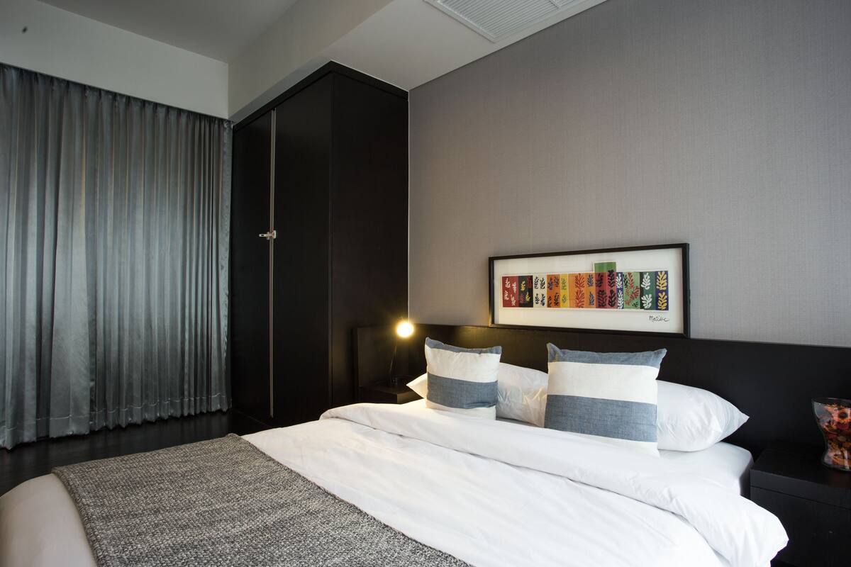 A modern bedroom with a large bed, a nightstand with a lamp, a wardrobe, and a framed artwork on the wall.