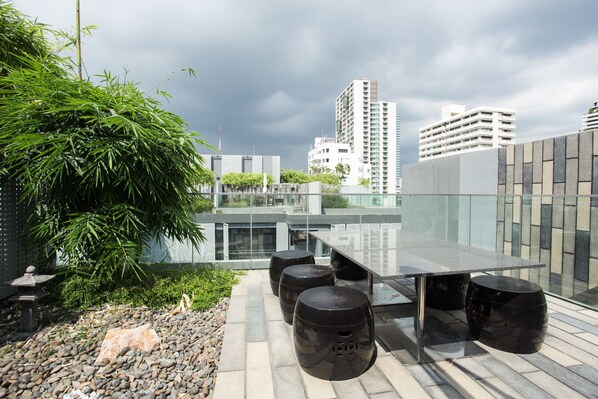 Eine Dachterrasse mit Glastisch und schwarzen Hocker, umgeben von Grünpflanzen und modernen Gebäuden.