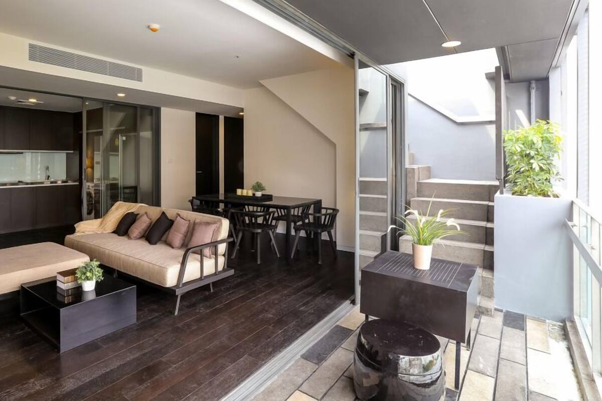 A modern interior with a sofa, dining table, and chairs, a kitchen area, and a staircase leading to an outdoor balcony.