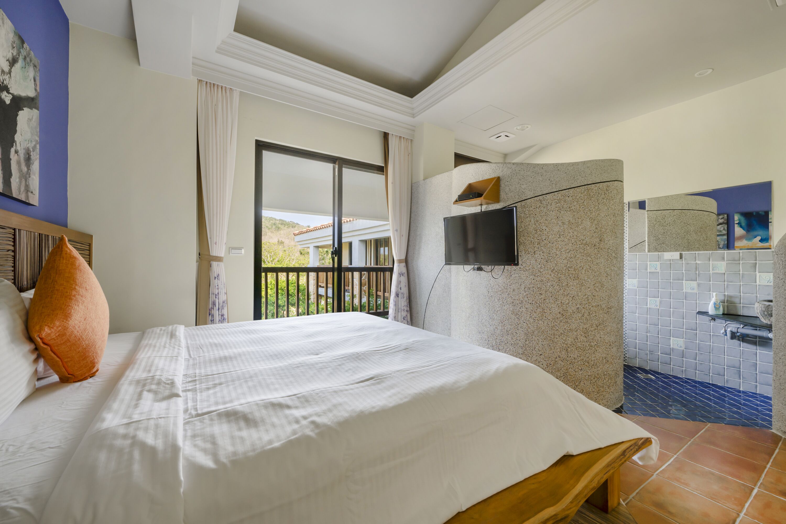 Une chambre d’hôtel dotée d’un grand lit, d’un téléviseur fixé à un mur en pierre, d’une douche et d’un balcon offrant une vue sur la verdure.