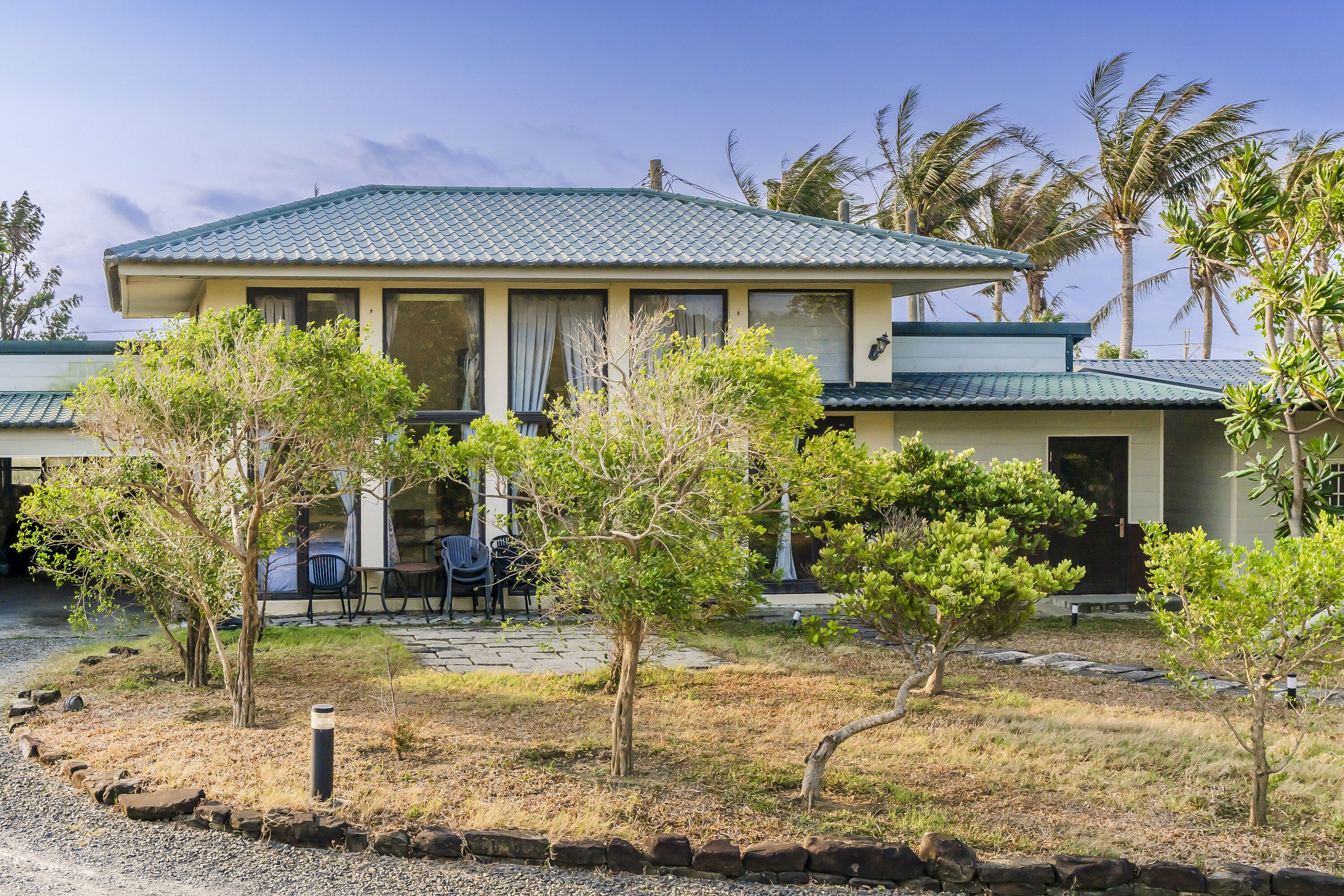 A two-story building with a tiled roof, a covered patio area with chairs, and a small garden with trees and shrubs.