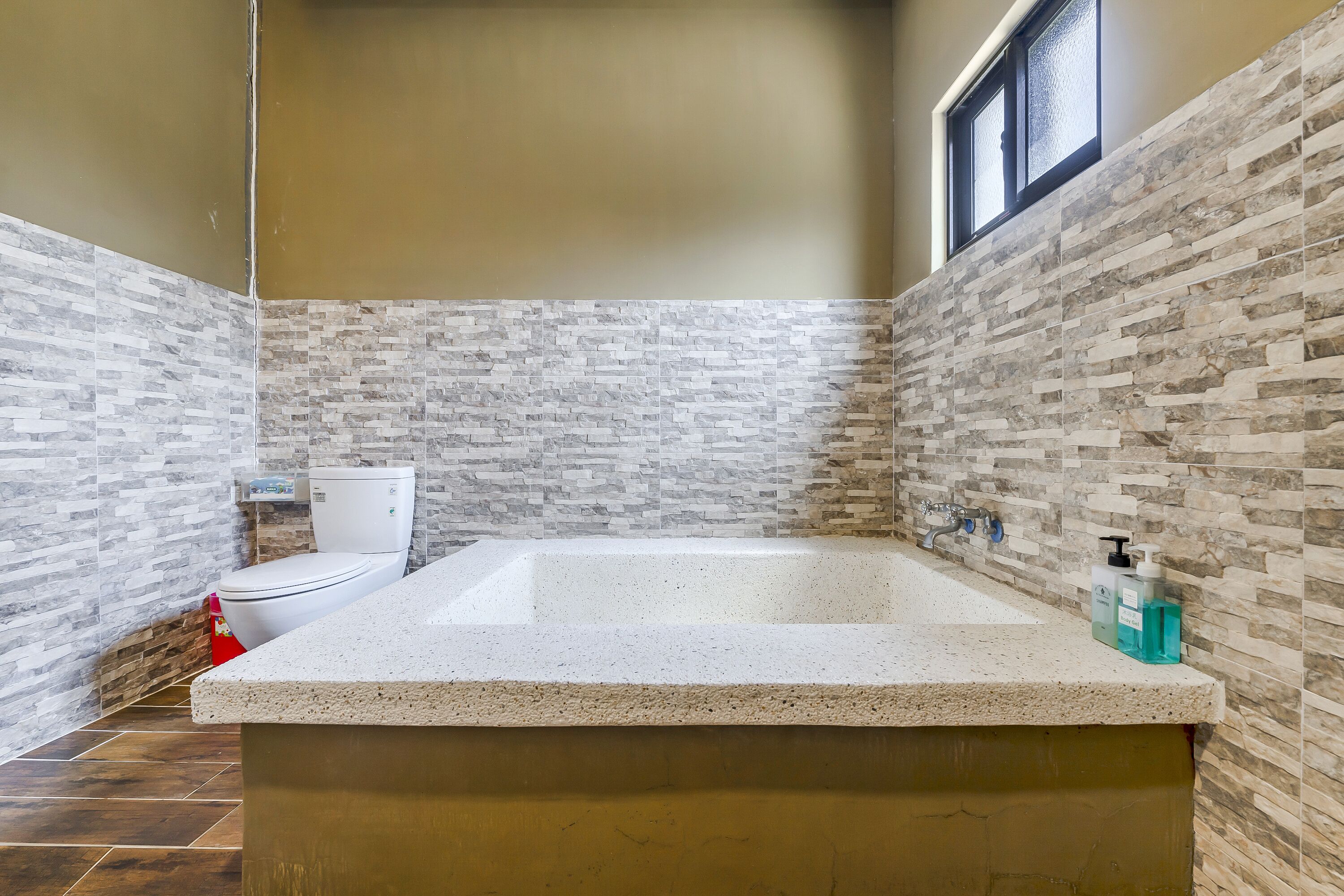 A bathroom with a stone mosaic wall, a white toilet, and a bathtub with soap dispensers.