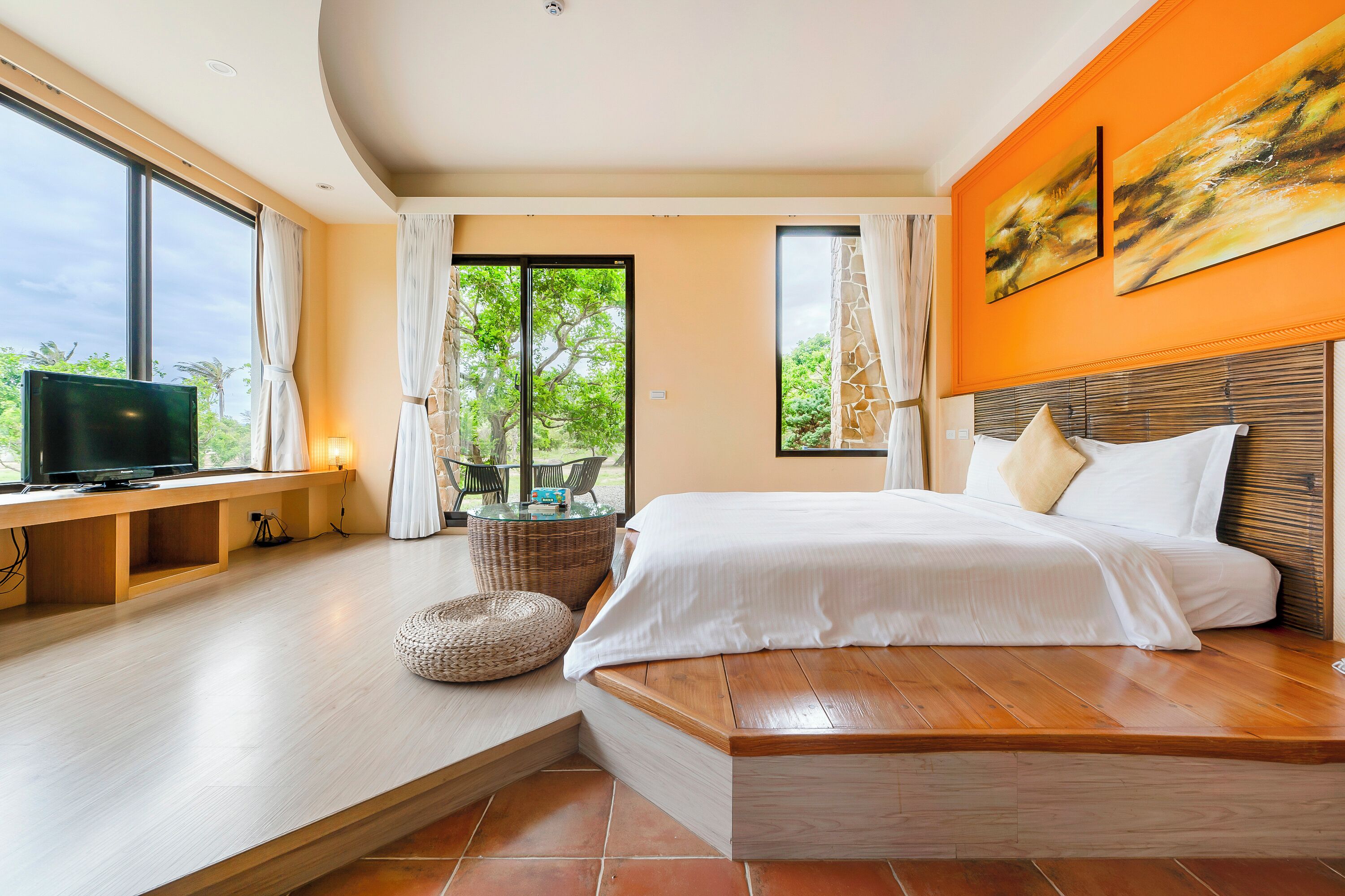 A modern bedroom with a large bed, a TV on a wooden console, a view of greenery through the windows, and a bright orange accent wall.