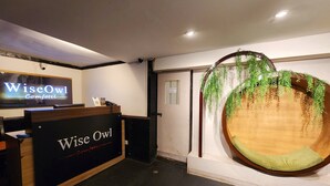 A reception area with a wooden desk labeled "Wise Owl Comfotel," a large screen displaying the same name, and a unique circular wooden bench with hanging greenery. - Wise Owl Comfotel (Kolkata)