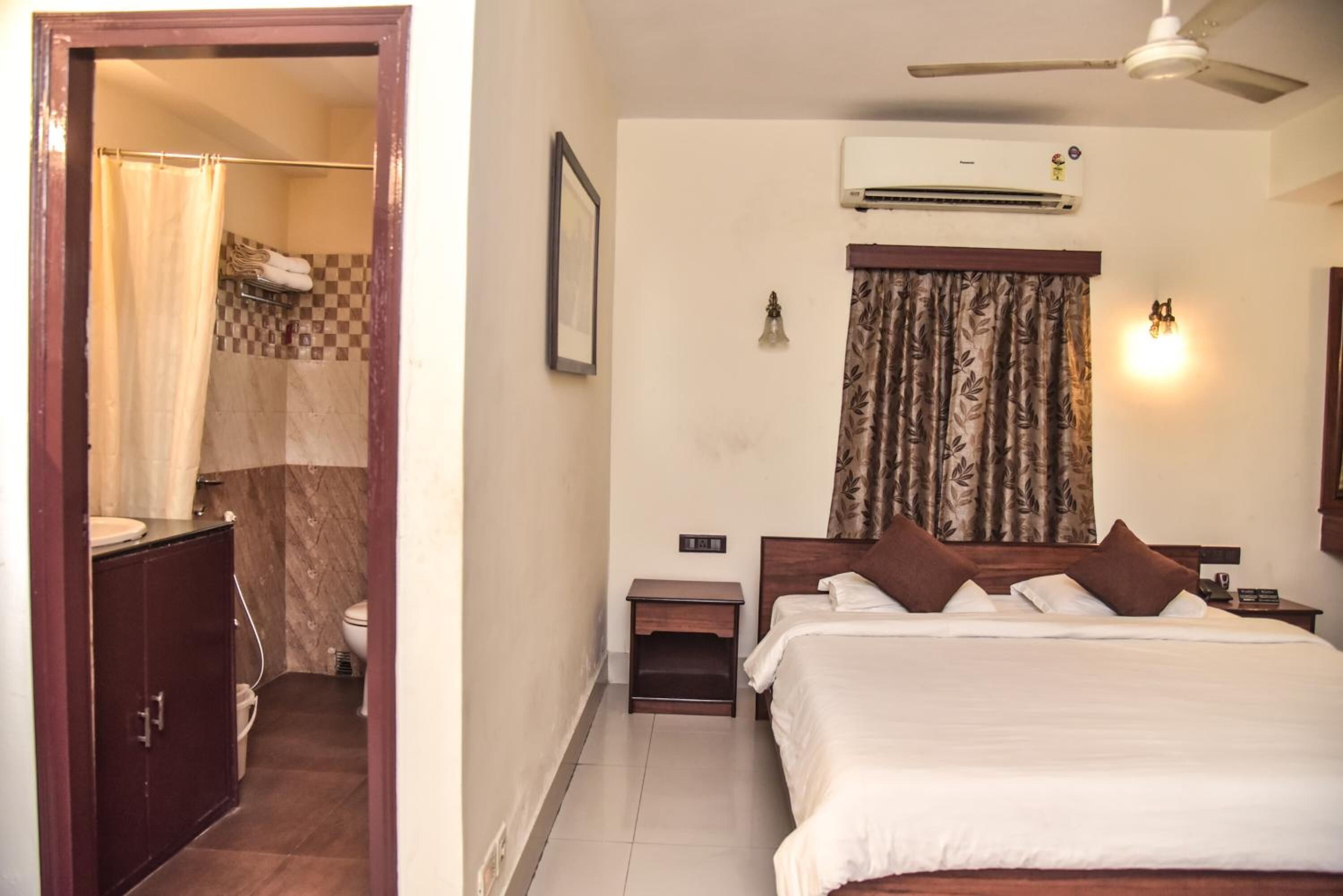 A hotel room with a bed, a nightstand, a small table, an air conditioning unit, a ceiling fan, a shower curtain, a toilet, and a cabinet.