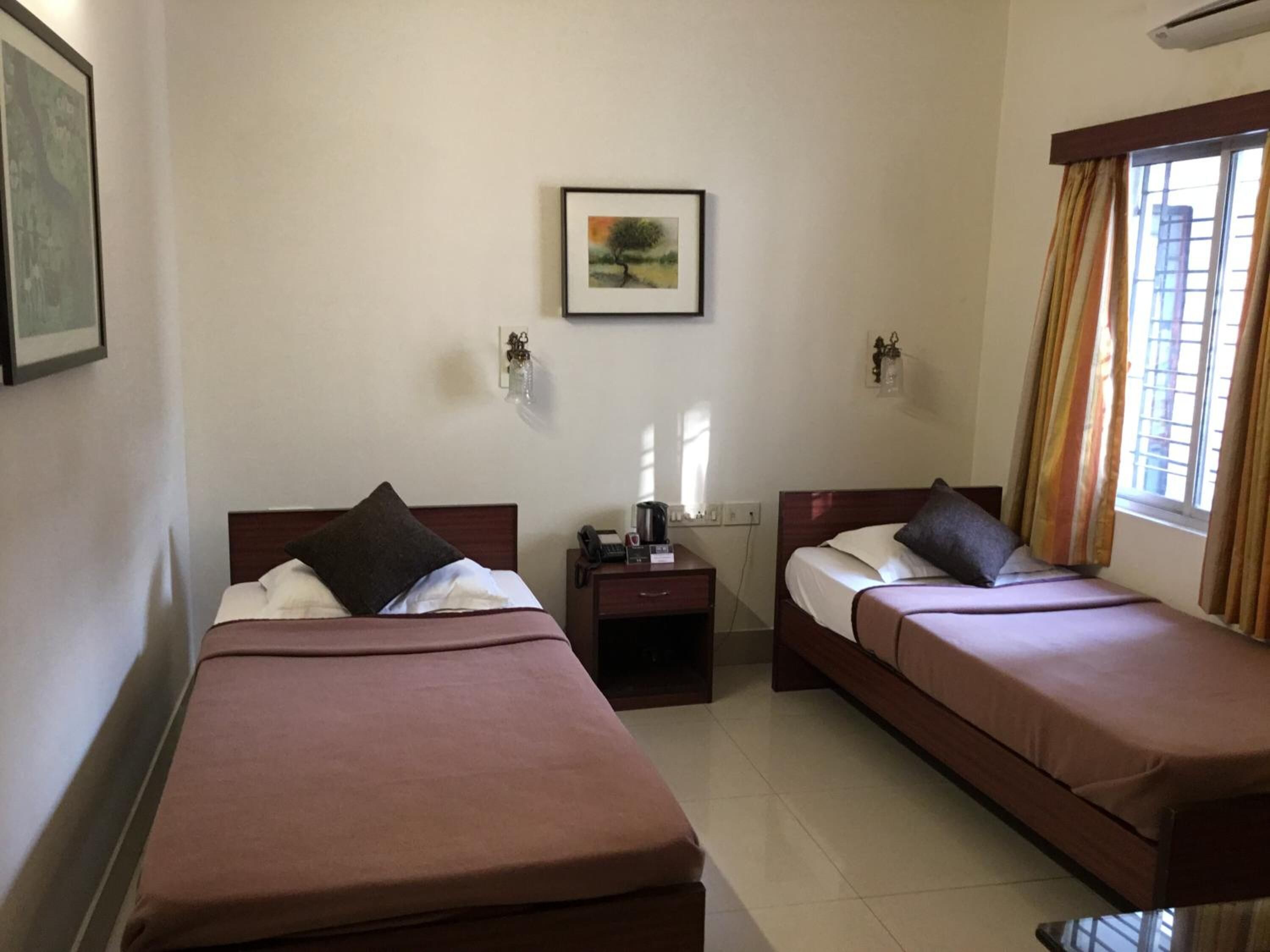 A hotel room with two single beds, a small table with a phone and a lamp, a picture on the wall, and a window with curtains.