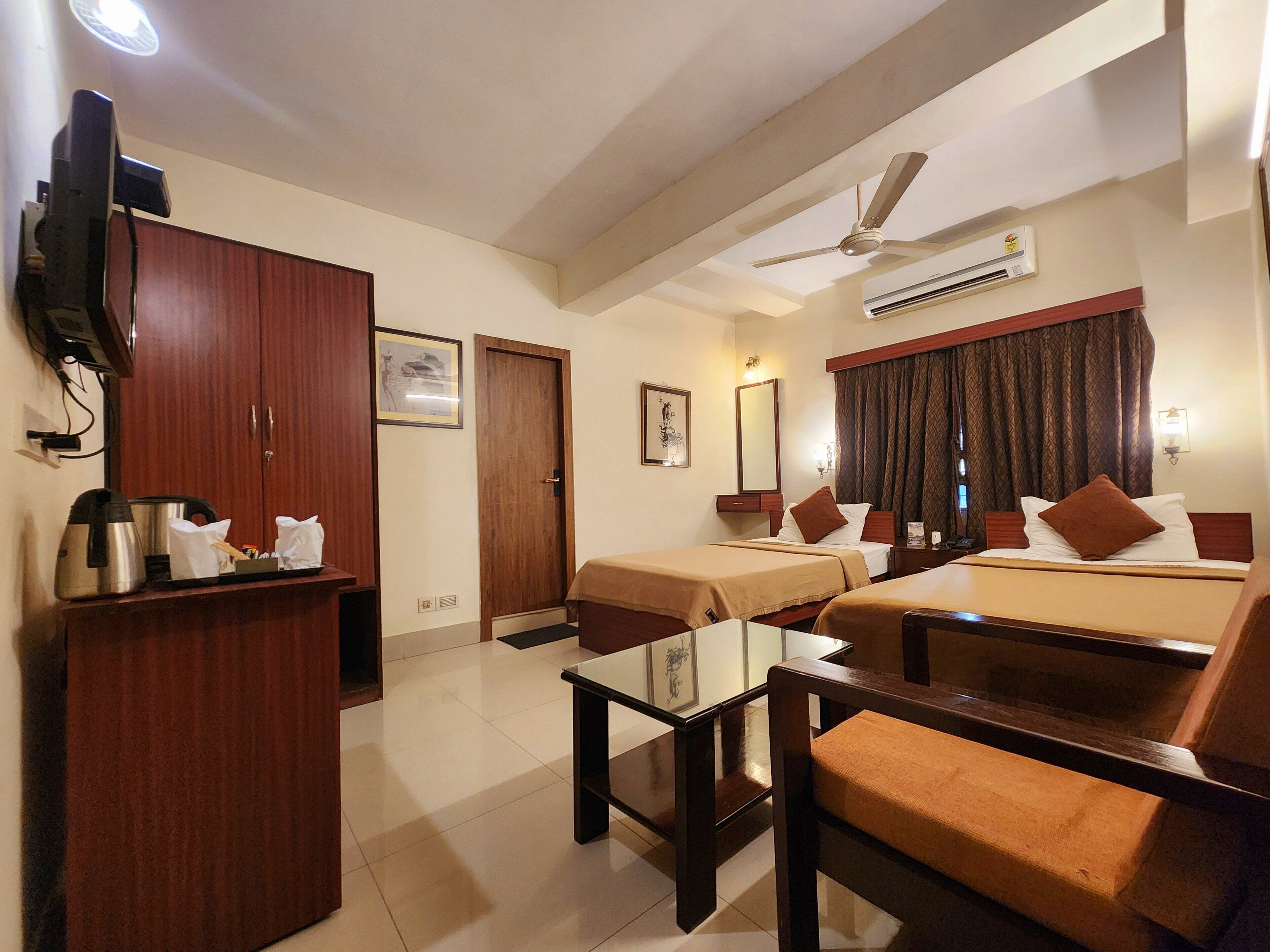 A hotel room with two single beds, a sofa bed, a wooden wardrobe, a small table, a ceiling fan, and an air conditioning unit.