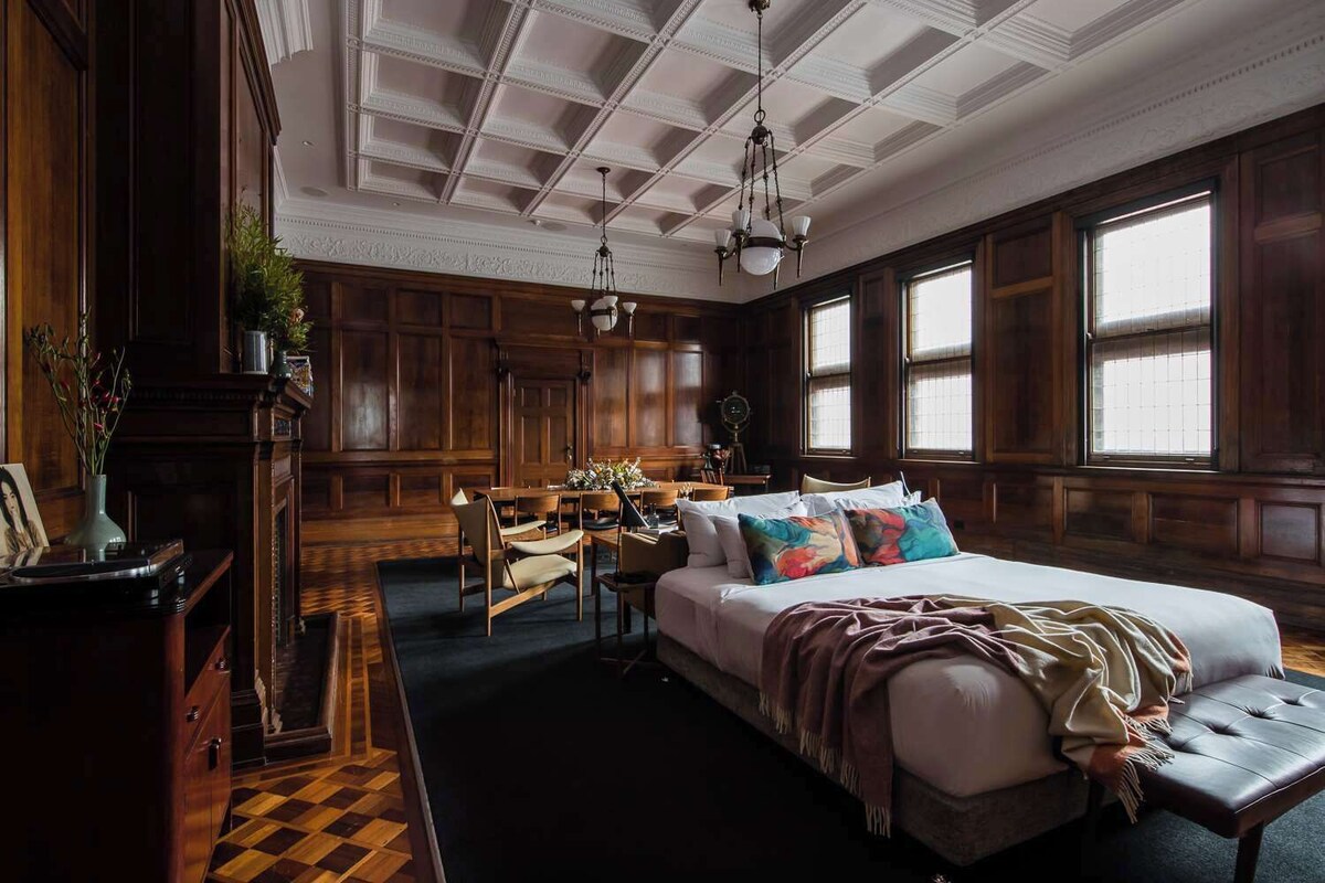 A room with a large bed, a desk, and a chair. There are windows and a chandelier.
