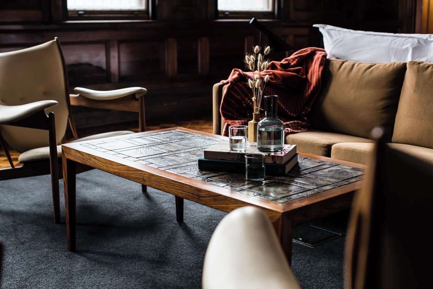 A cozy room with a wooden coffee table, a book, a glass of water, and a vase with dried flowers. There is a brown sofa and a chair in the background.
