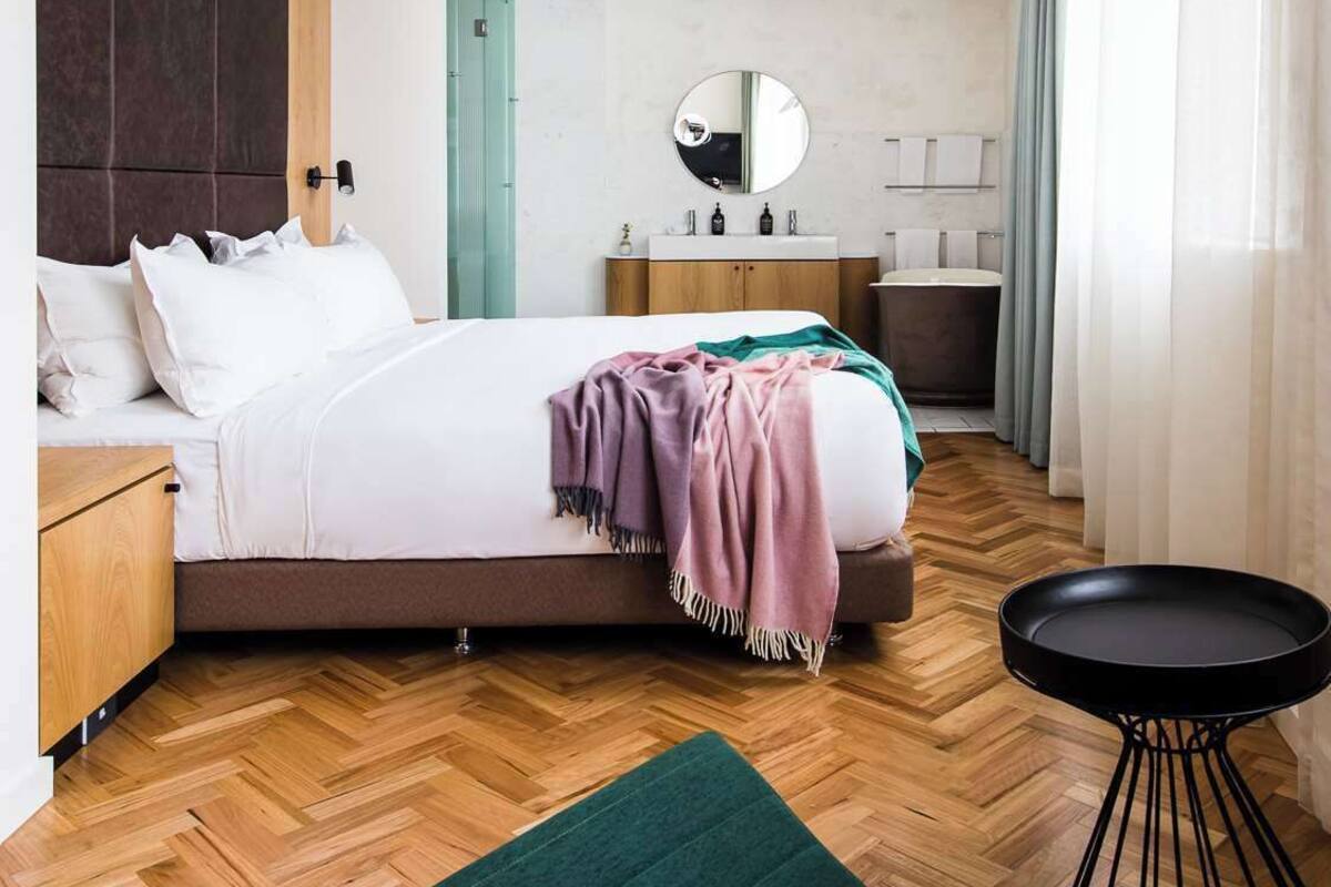 Un dormitorio moderno con cama, mesitas de noche, un espejo redondo, una pequeña mesa redonda y un otoman verde.