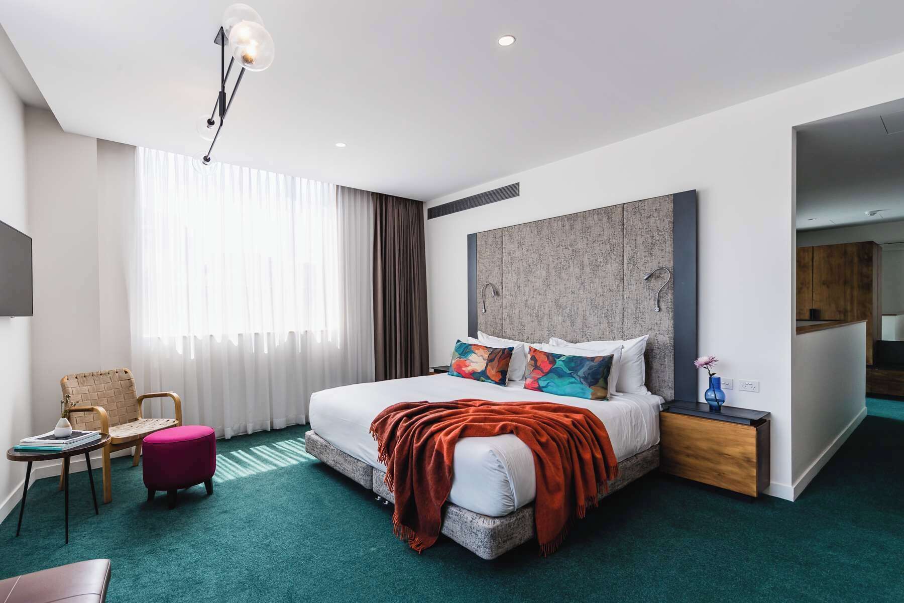 A modern hotel room with a large bed, a wooden headboard, a small bedside table, a chair, and a large window with sheer curtains.
