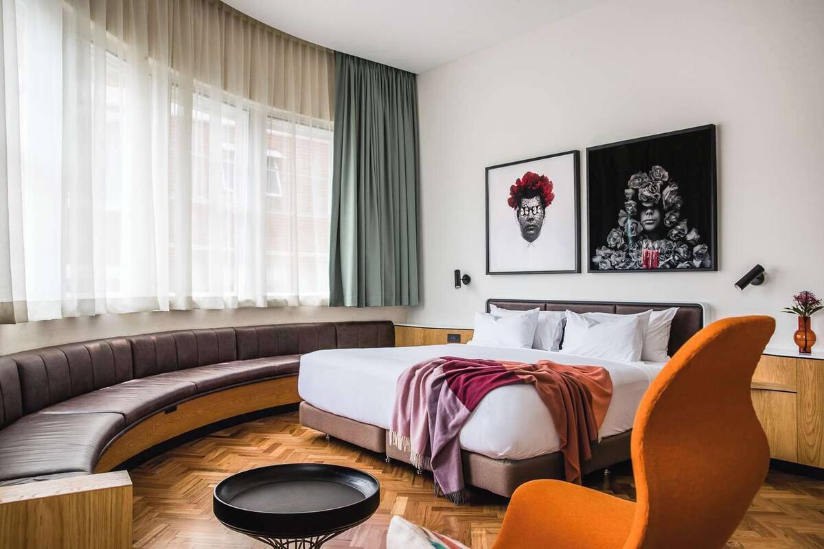 Una habitación de hotel moderna con una zona de estar curva, una cama con una colcha roja y dos obras de arte enmarcadas en la pared.