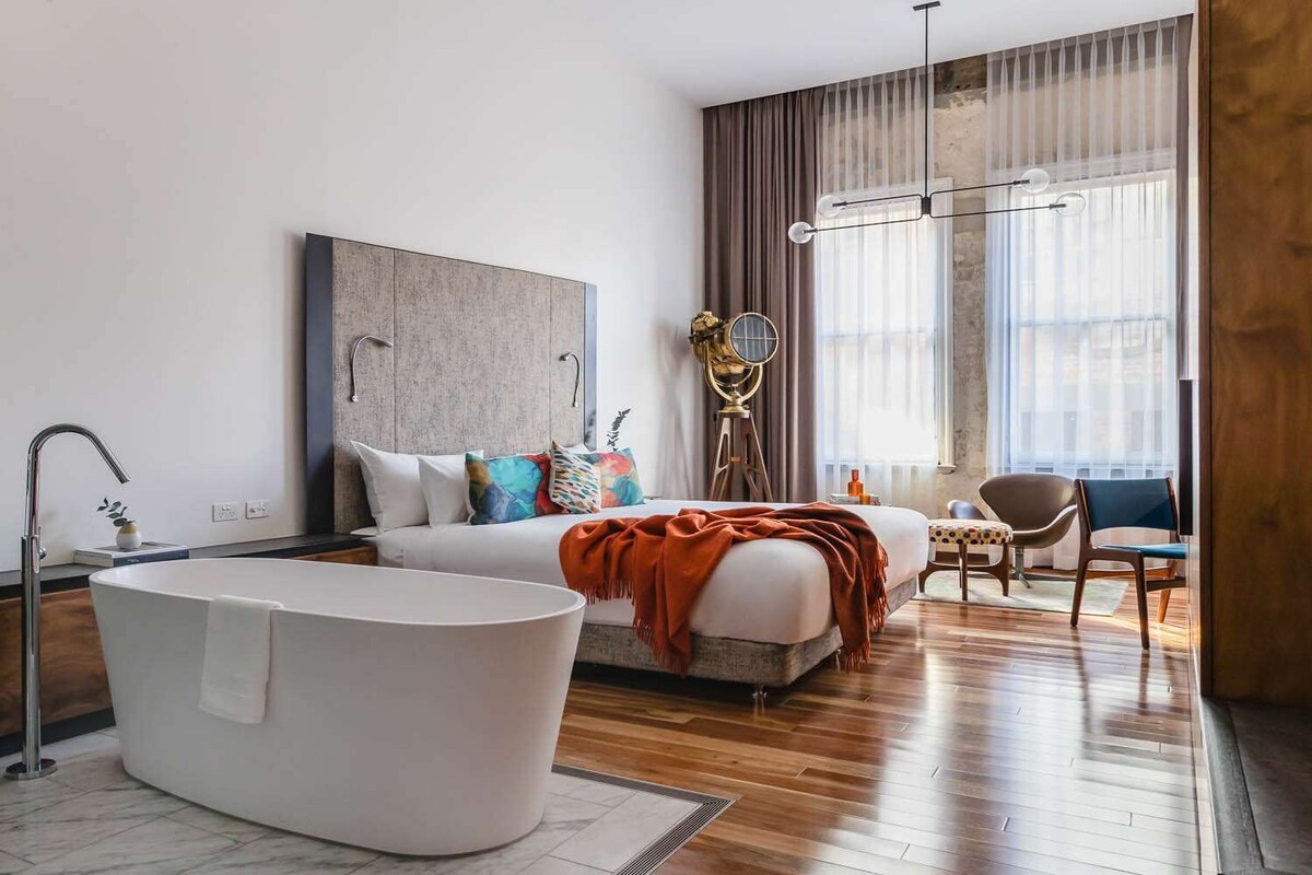 A modern hotel room with a large bed, a freestanding bathtub, and a large window with sheer curtains.