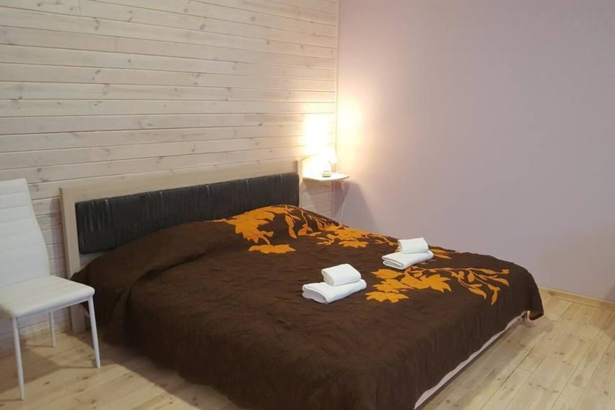 A bedroom with a bed, a nightstand with a lamp, a white chair, and a small table.
