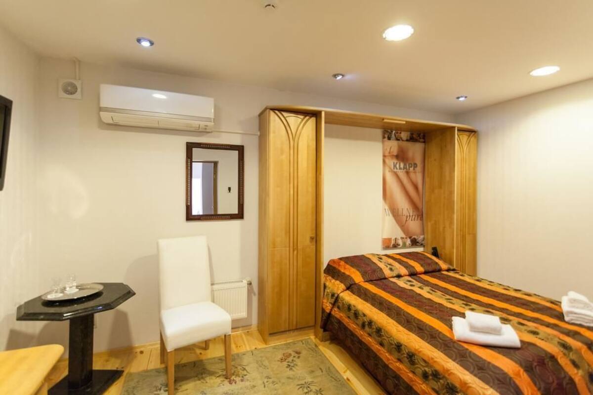 A hotel room with a bed, a chair, a small table, an air conditioning unit, and a wooden wardrobe.