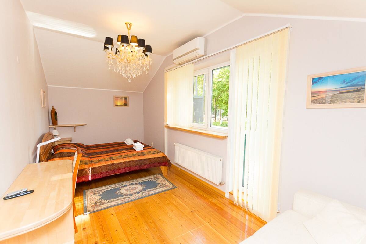 A bedroom with a bed, a desk, a chandelier, a window with curtains, and a radiator.