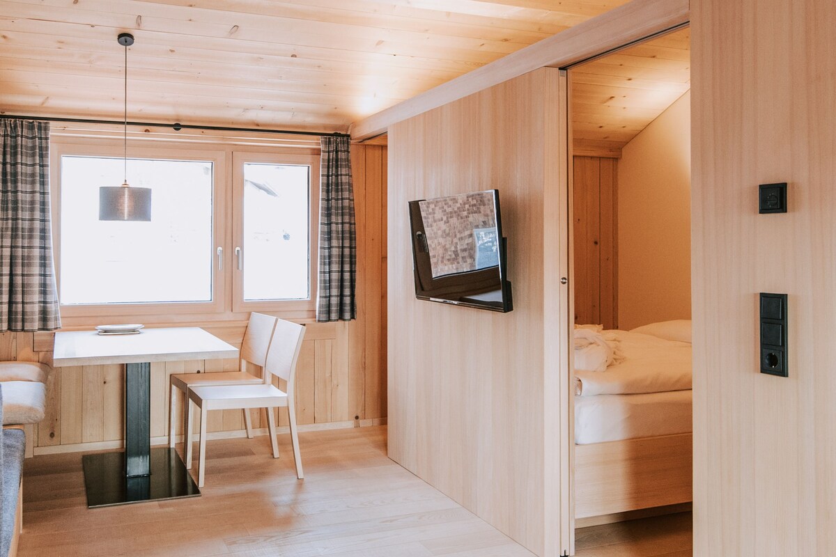 Quarto com teto de madeira, uma cama, televisão fixada na parede, janela com cortinas e mesa com cadeiras.