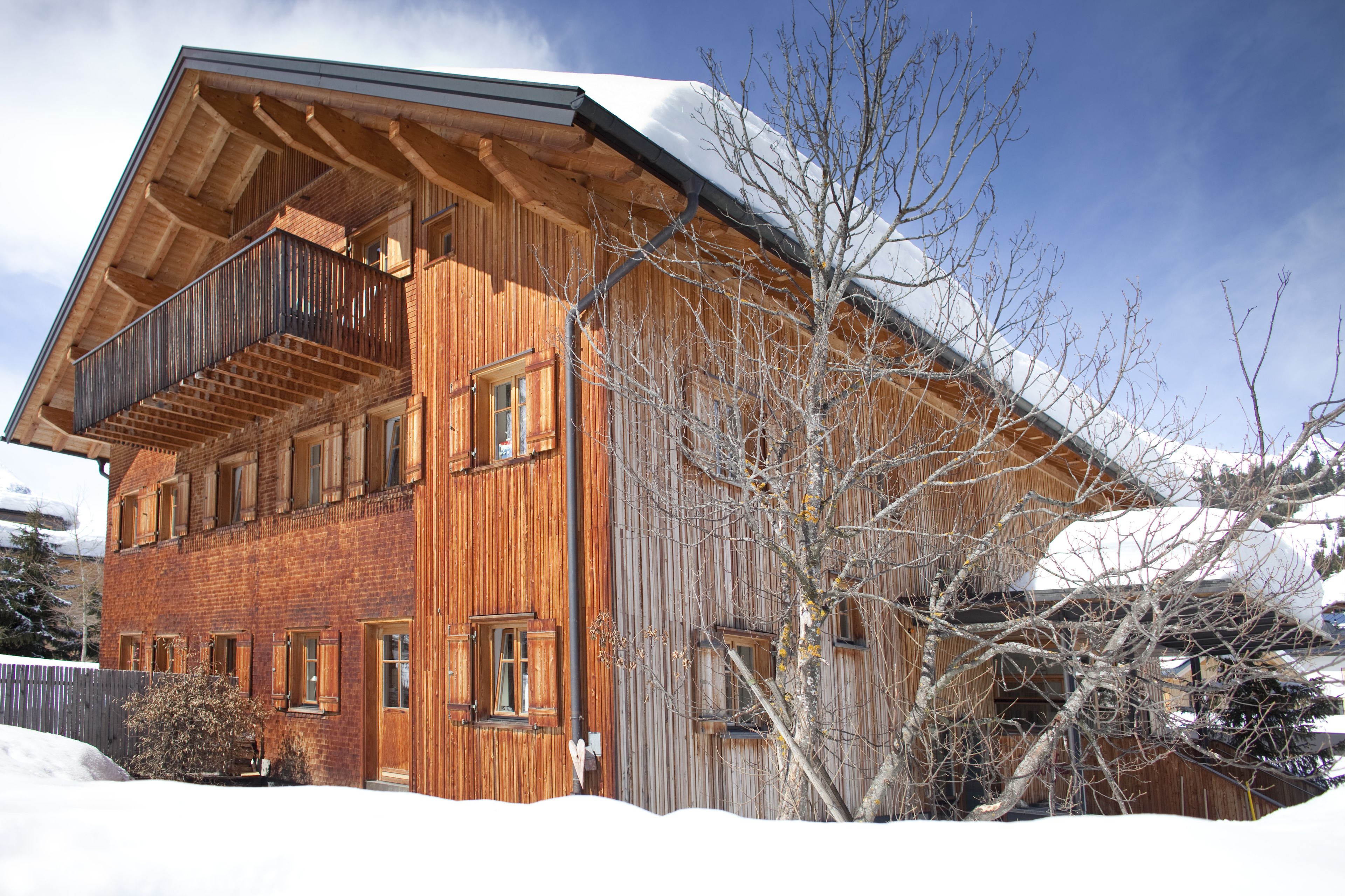 A wooden chalet with a balcony, large windows, and a snow-covered roof.
