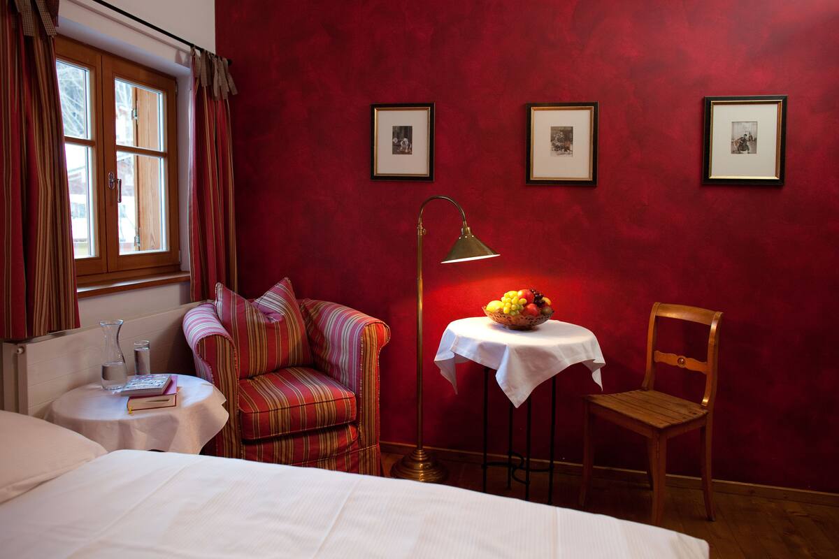 A hotel room with a bed, a striped armchair, a wooden chair, a small table with a fruit bowl, a floor lamp, and framed pictures on the wall.
