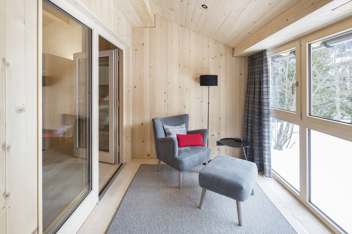 A modern room with a wooden interior, a grey armchair with a red pillow, a grey ottoman, a floor lamp, and a large window with curtains.