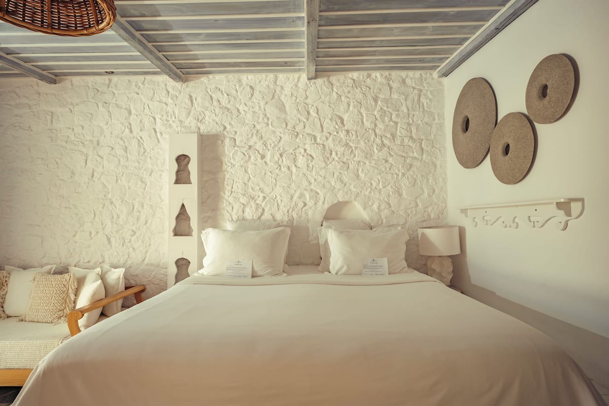 Een slaapkamer met een wand met een stenen textuur, een bed met witte beddengoed, een houten bankje en een wandplank met decoratieve objecten.