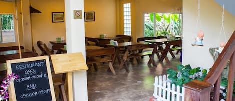 Interior de un restaurante con mesas y sillas de madera, un cartel de pizarra con el horario de atención y una escalera con valla de piquetes blancos.