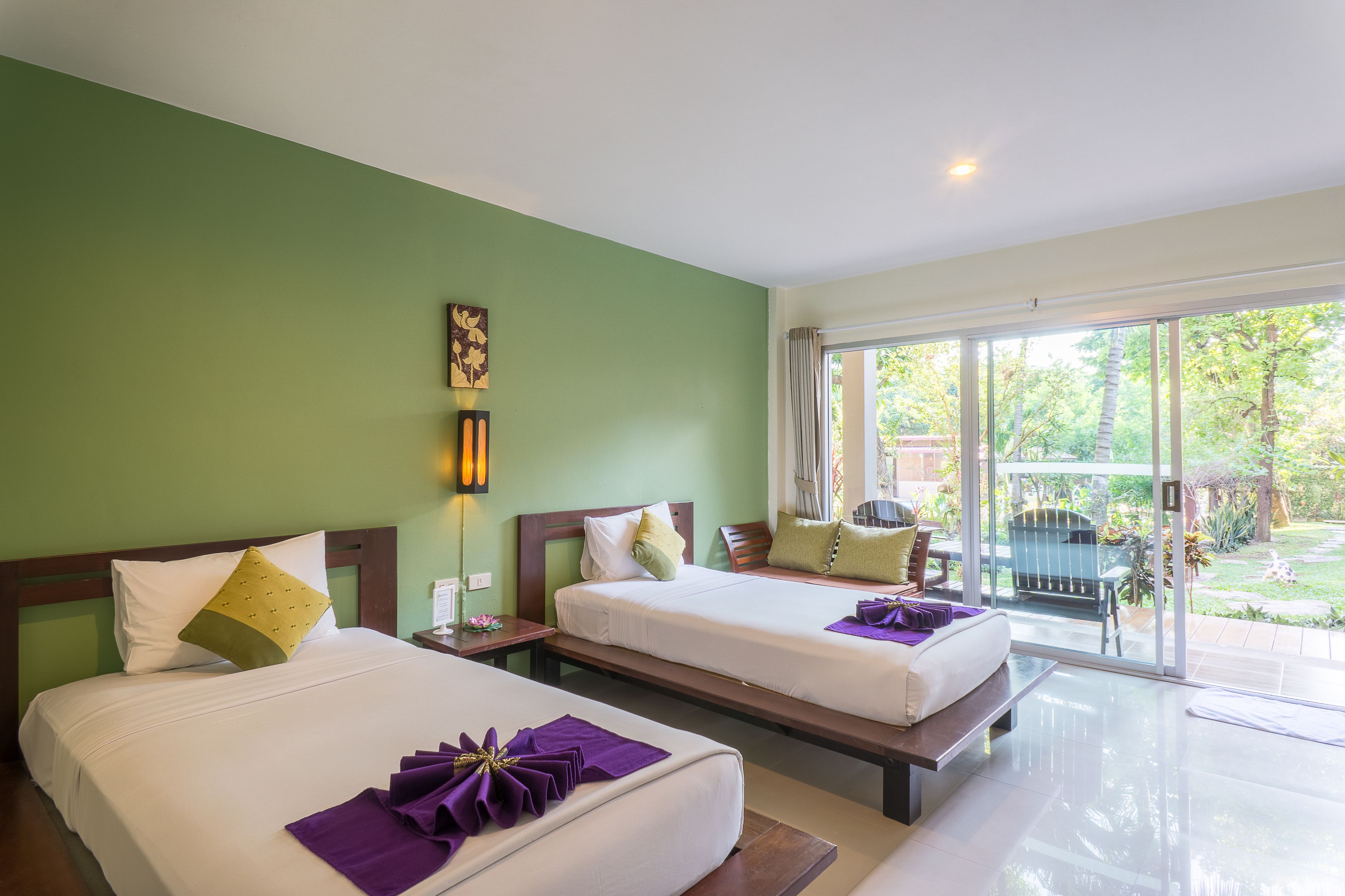 A hotel room with two beds, a green wall, a window with a view of the garden, and a small table with a chair.