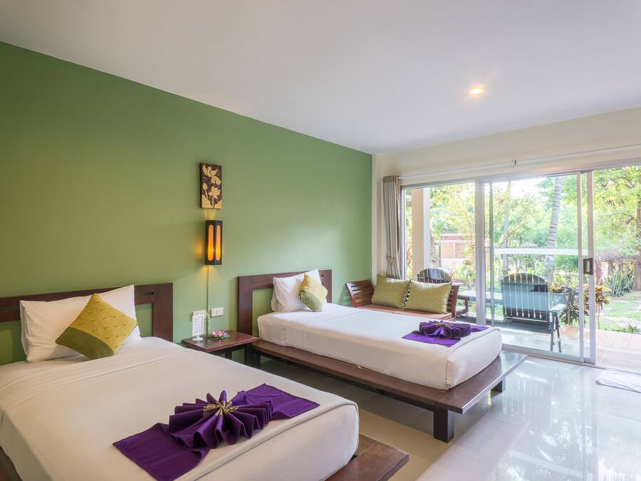 A hotel room with two beds, a green wall, a window with a view of the garden, and a small table with a chair.
