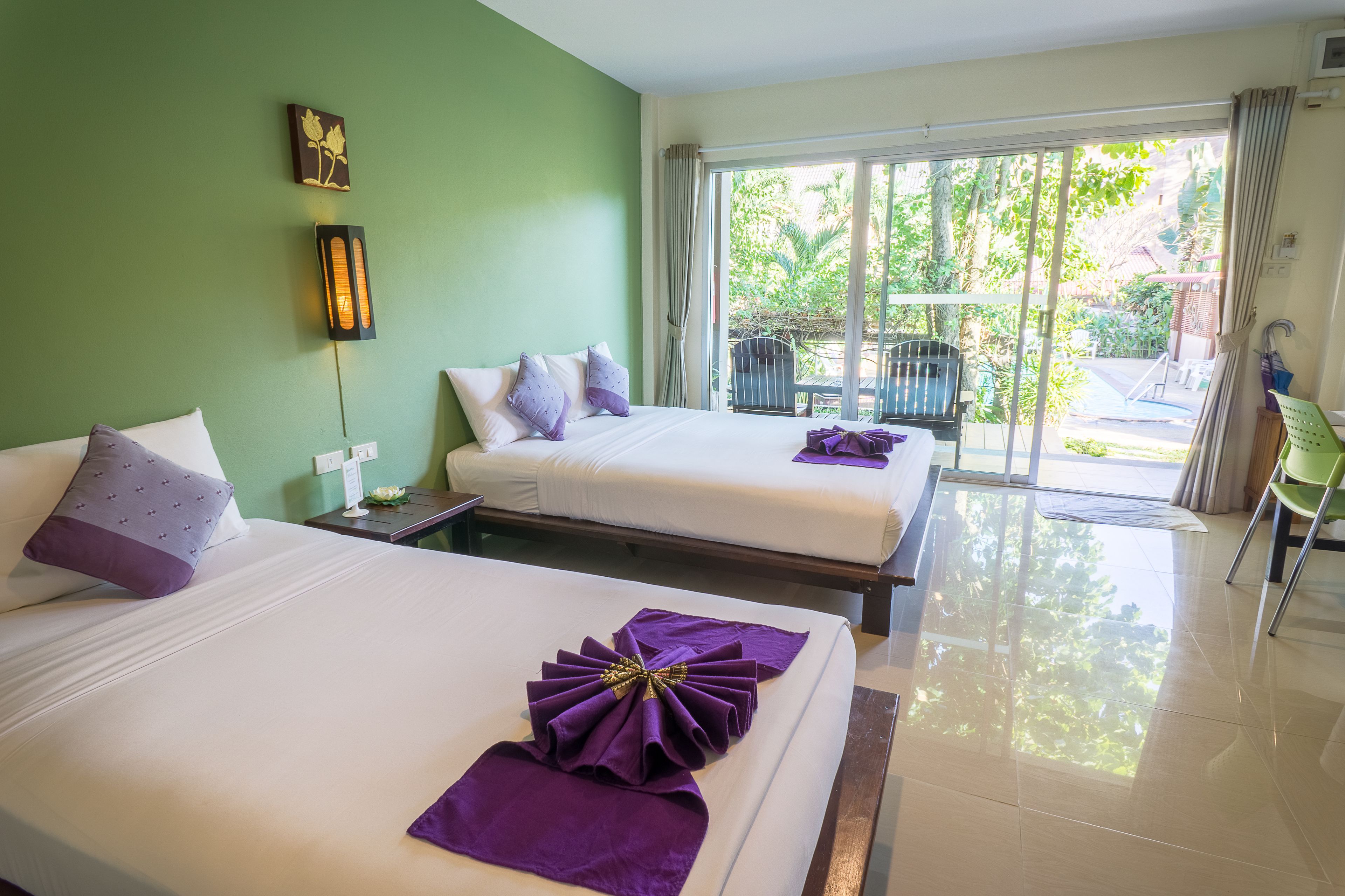 Habitación de hotel con dos camas, una pared verde, una puerta corredera de vidrio a un balcón y vistas a la vegetación.