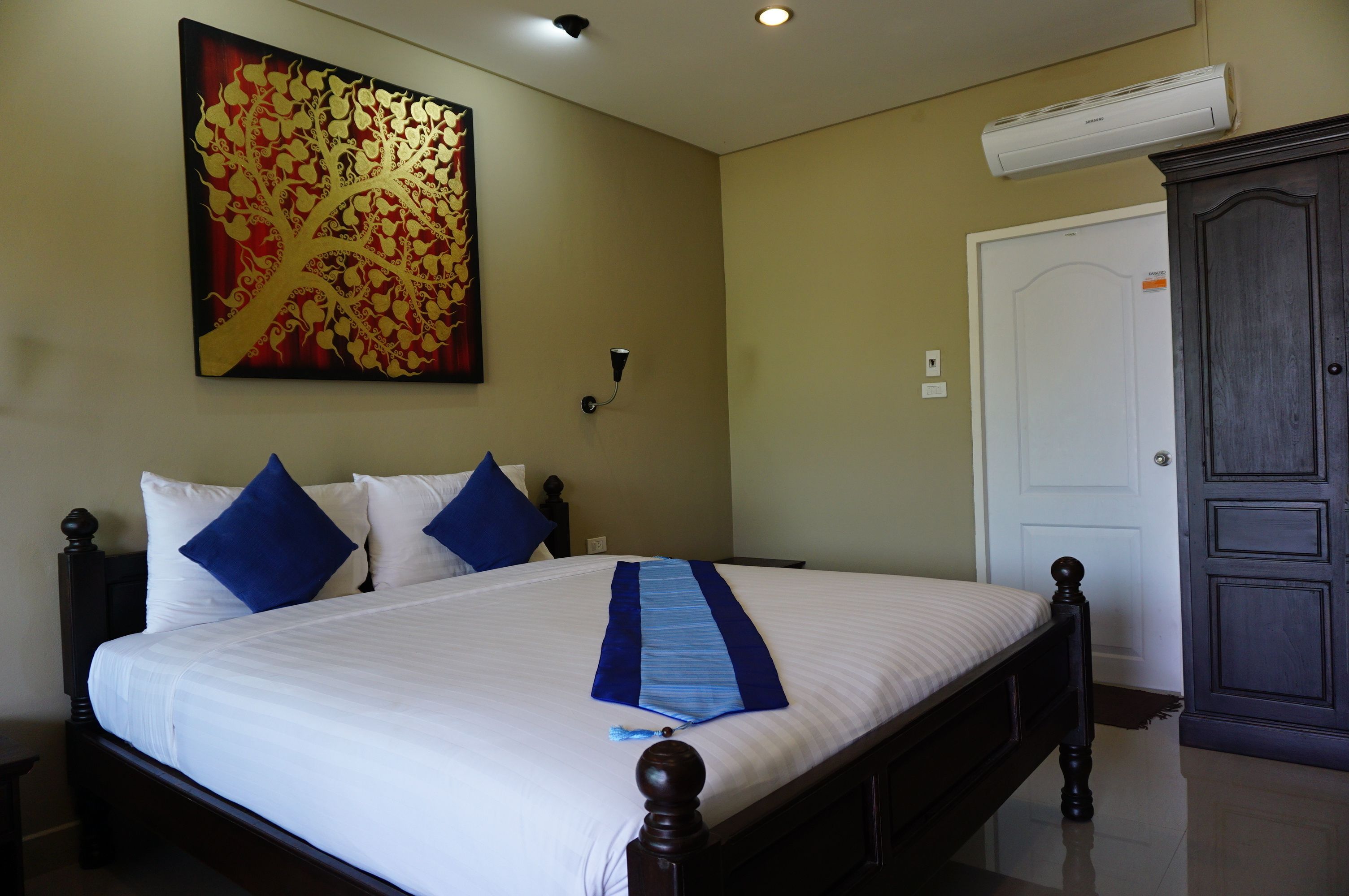 A neatly made bed with blue pillows, a decorative blue and white runner, a large abstract painting on the wall, a dark wooden wardrobe, and a white door.