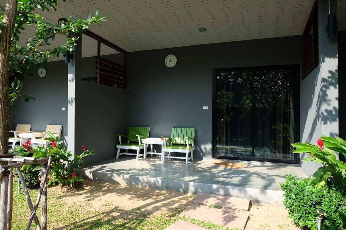 Un espacio al aire libre cubierto con dos bancos verdes y una mesa blanca, rodeado de follaje y un árbol.