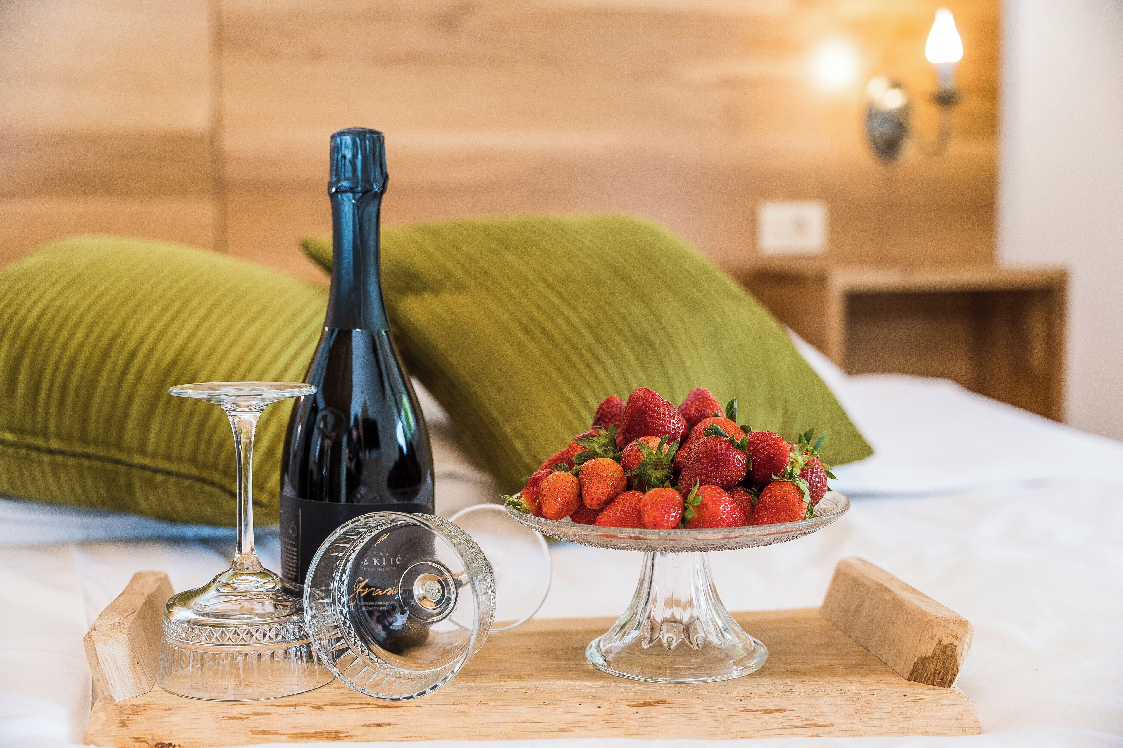 A bed with a wooden headboard, a bottle of wine, a wine glass, and a bowl of strawberries on a wooden tray.