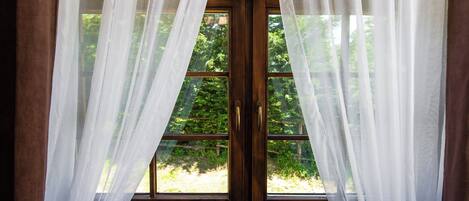 A room with a large window covered by sheer white curtains, offering a view of greenery outside.