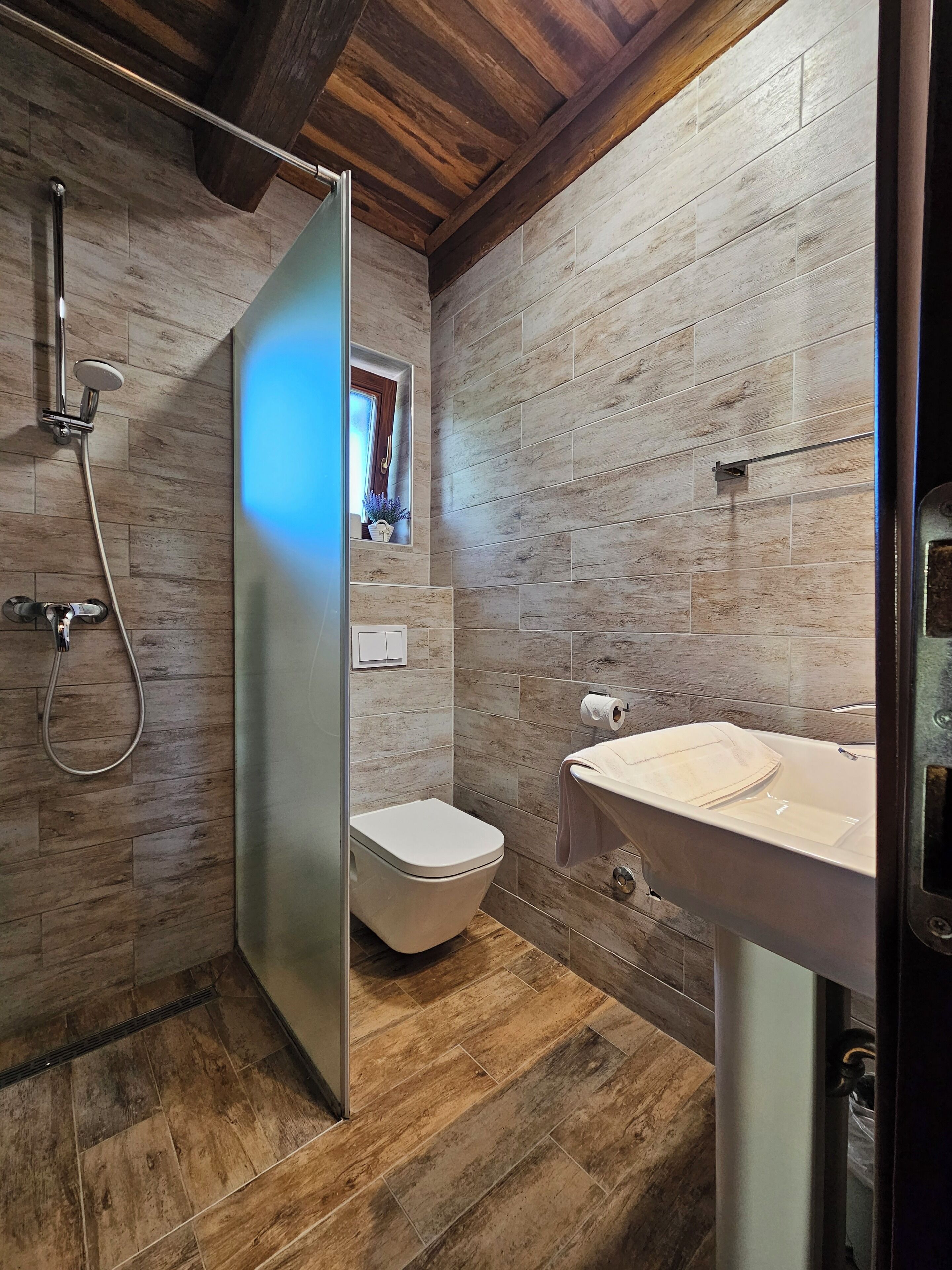 A modern bathroom with a wooden floor, a shower with a glass door, a toilet, and a sink with a towel hanging on it.