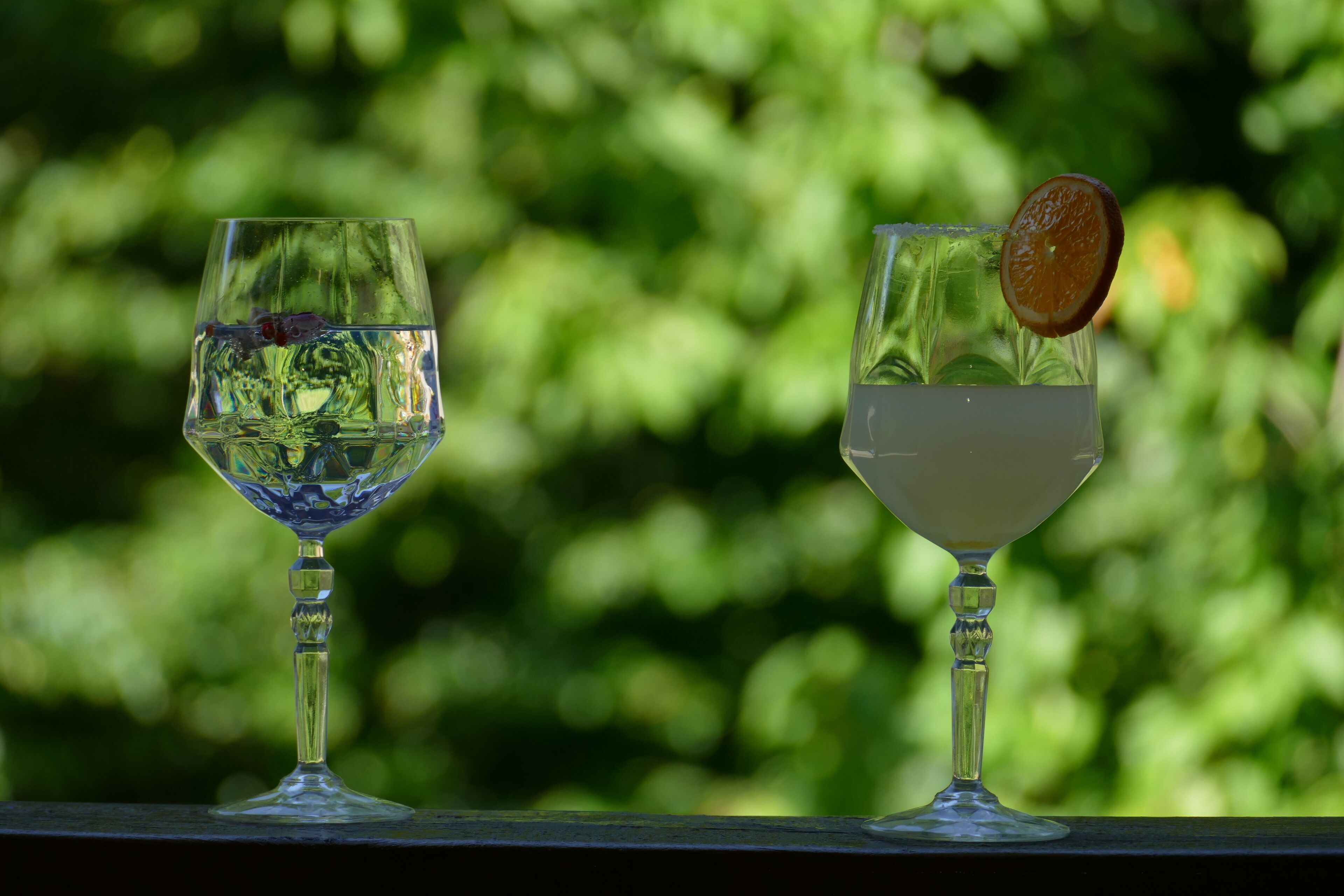 Two clear glasses of water on a balcony with a blurred green background.