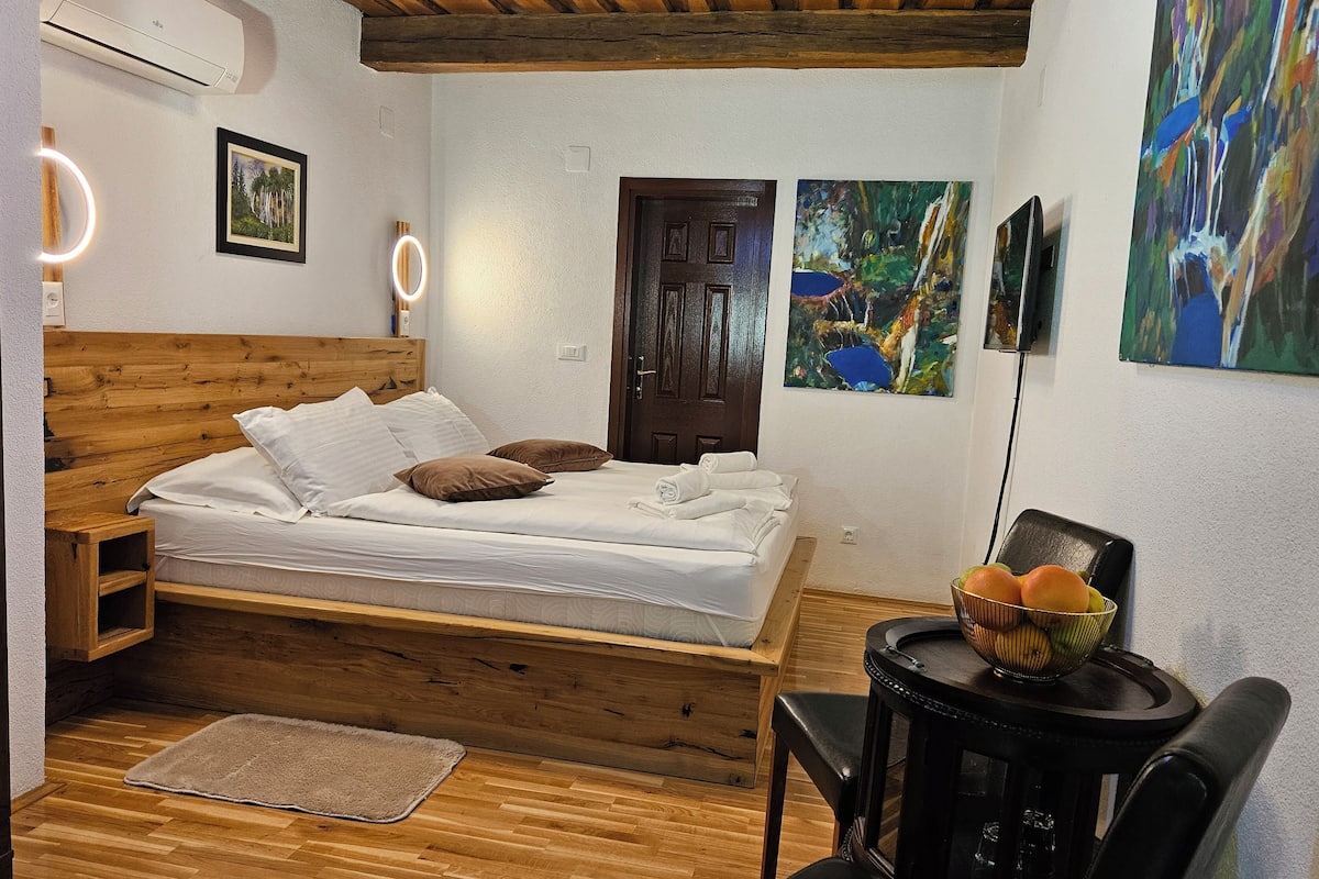 A wooden bed with white linens, a wooden nightstand, a round table with a fruit bowl, and two paintings on the wall.