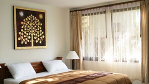 Una cama bien hecha con colcha marrón, un reloj en la pared y una ventana con cortinas traslúcidas.