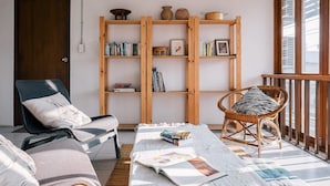 A cozy living room with a wooden bookshelf, a comfortable chair, a sofa, and a large window. - Happynest Inspiring Hostel (Chiang Rai)