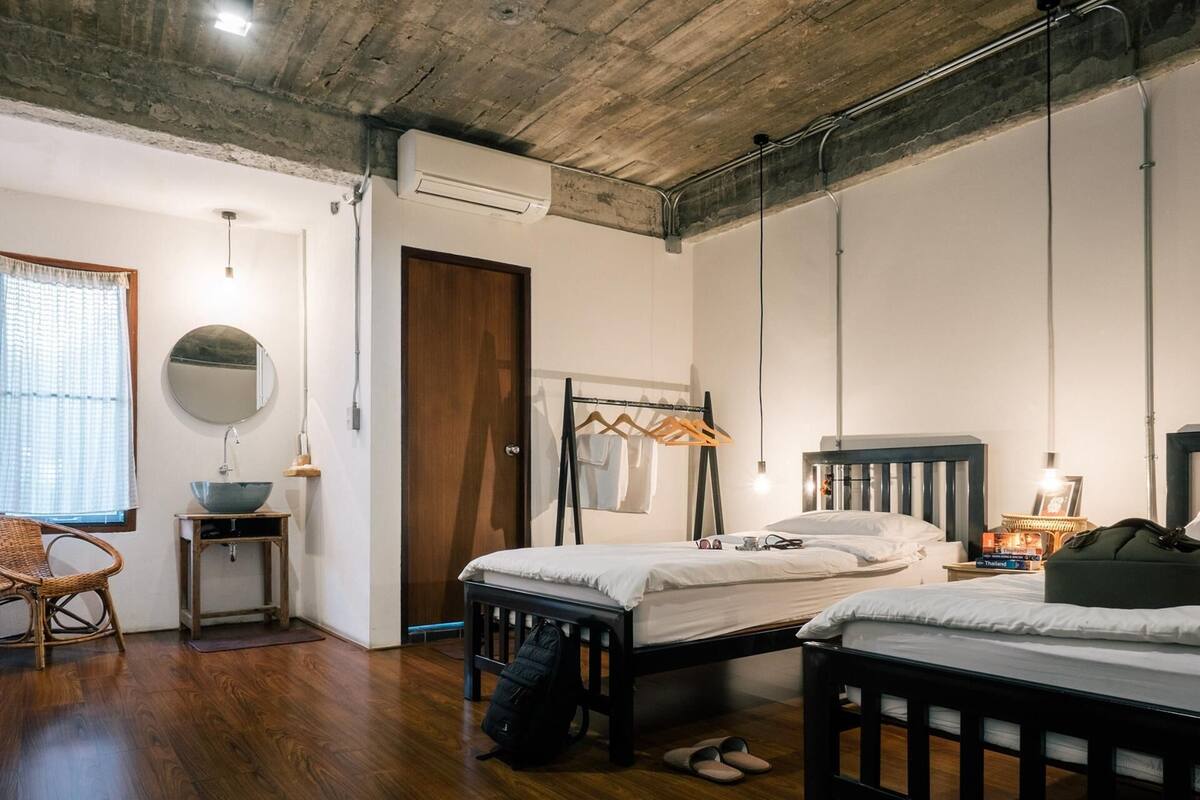 A bedroom with a wooden floor, a bed, a bedside table with a bowl, a hanging light, a wardrobe, and a window with a curtain.