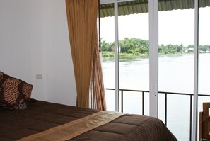 A bedroom with a large window overlooking a river, a bed with a brown bedspread, and a curtain. - Tara Raft Kanchanaburi (Kanchanaburi)