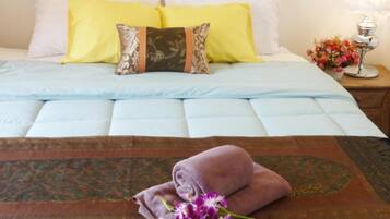 Una cama bien hecha con colcha azul, dos almohadas (una amarilla y otra marrón), un plaid decorativo marrón y una toalla púrpura.