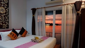 A hotel room with a bed, pillows, a pink blanket, and a view of a sunset over water. - Tara Raft Kanchanaburi (Kanchanaburi)
