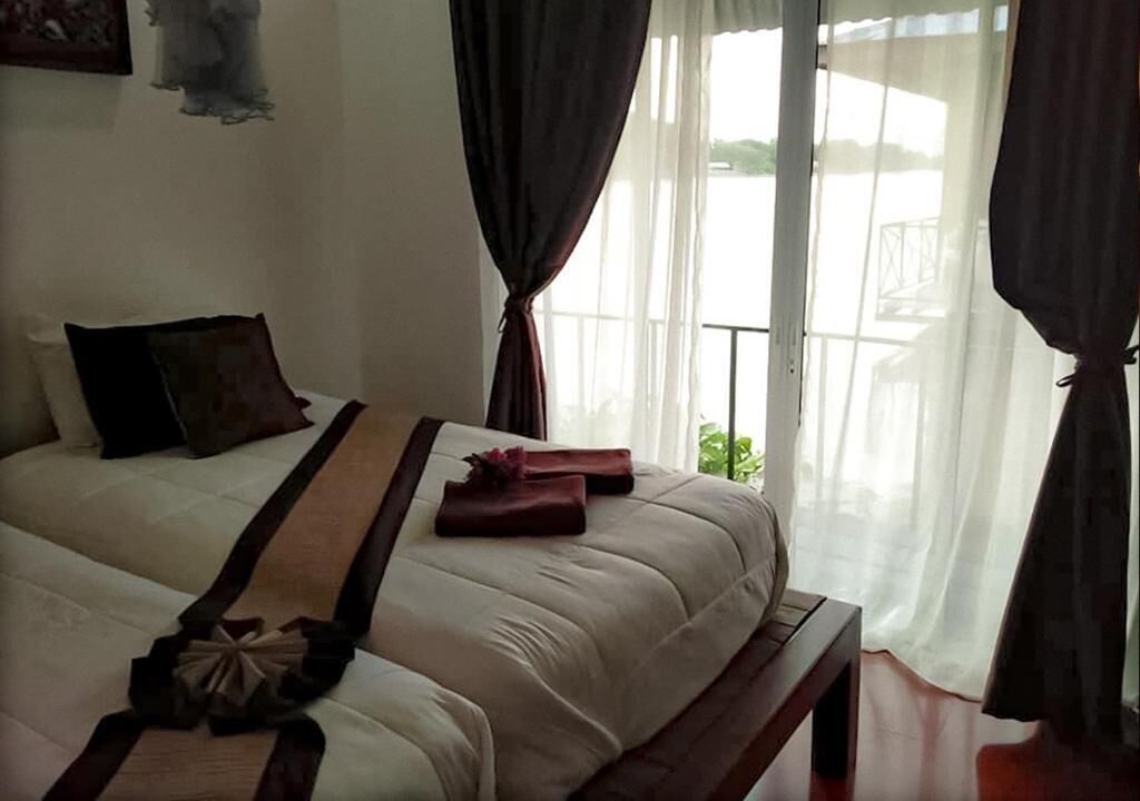 Ein ordentlich bezogenes Bett mit Kissen und Decke, ein Nachttisch und Blick auf einen Balkon durch transparente Vorhänge.