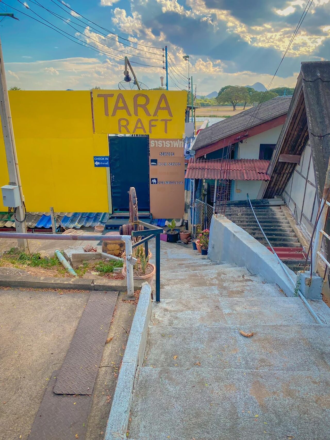 Eine Treppe führt zu einem Gebäude, dessen Wand gelb ist und auf der Beschriftung "TARA RAFT" zu lesen ist. Ein Schild zeigt die Optionen "CHECK IN" und "SERVICE" an.
