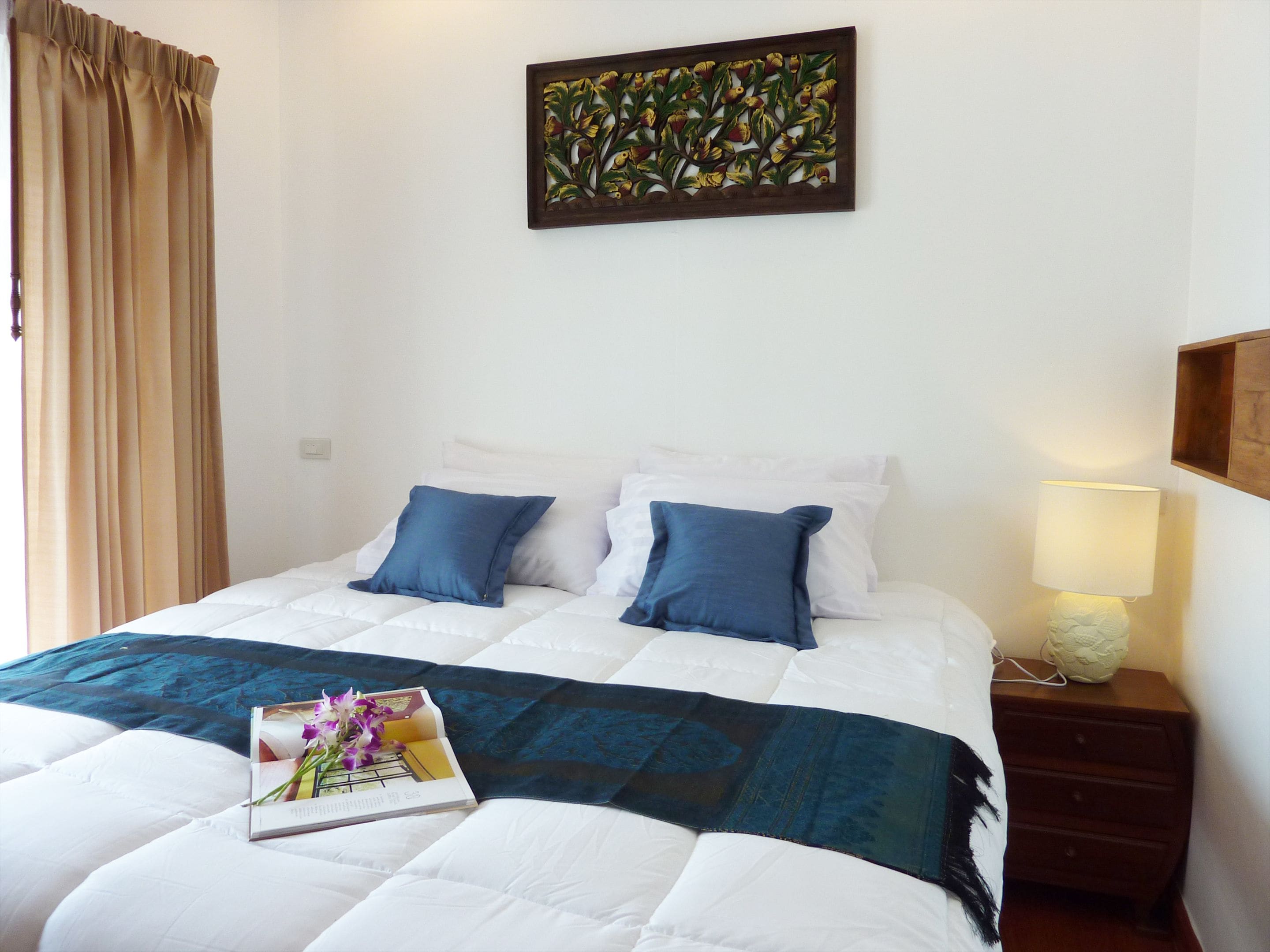 A neatly made bed with white and blue bedding, a decorative pillow, a book with flowers, and a wall art piece.