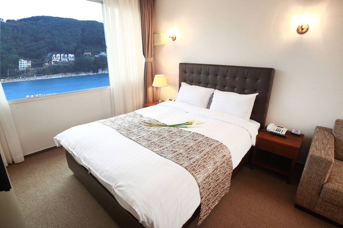 A hotel room with a large bed, a nightstand with a lamp, a chair, and a view of a body of water and buildings.