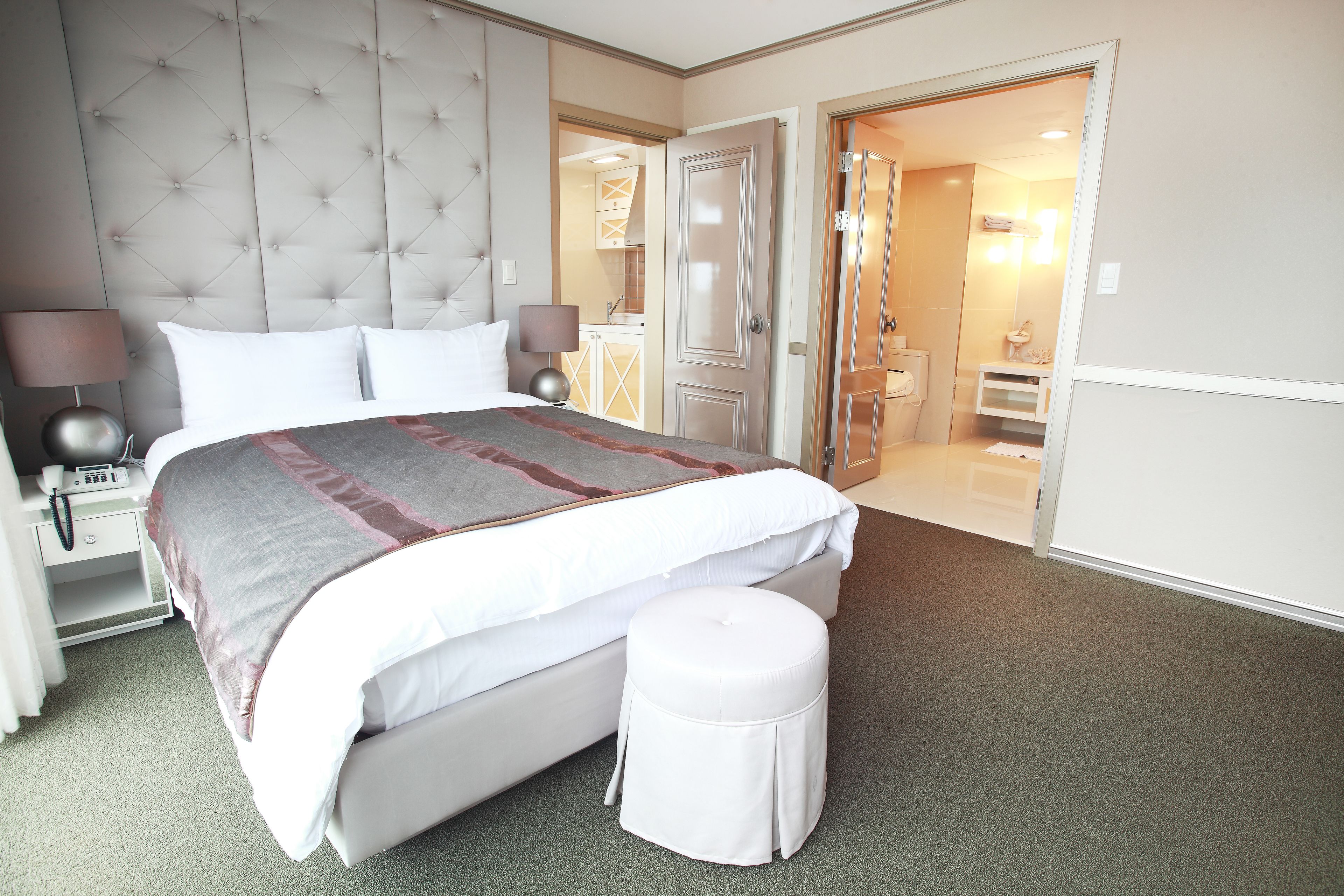 A hotel room with a large bed, a bedside table with a lamp, a small stool, and a door leading to a bathroom.