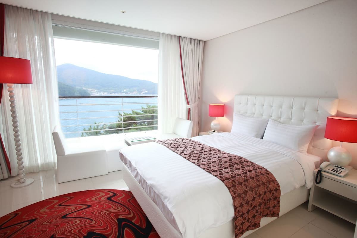 A hotel room with a large bed, two bedside lamps, a telephone, a window with curtains, and a view of the sea.