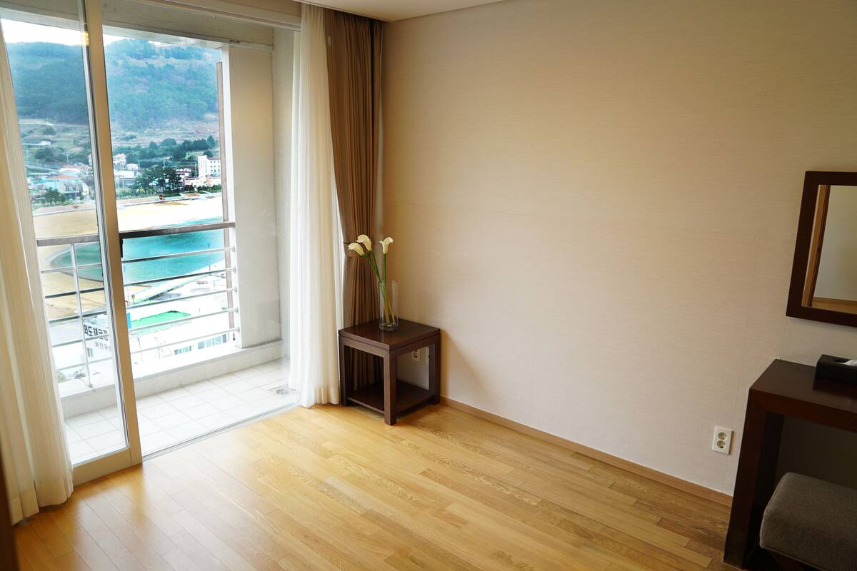 A room with a wooden floor, a small table with a vase of flowers, a view of a beach and mountains through a large window, and a chair.